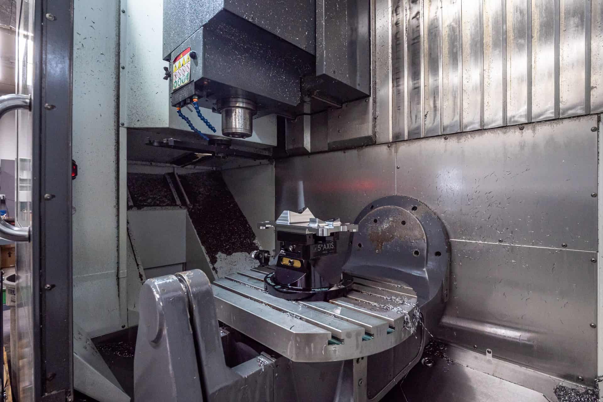 A CNC milling machine with a metal workpiece secured in a vise on the rotating table, surrounded by metal shavings inside the enclosure.